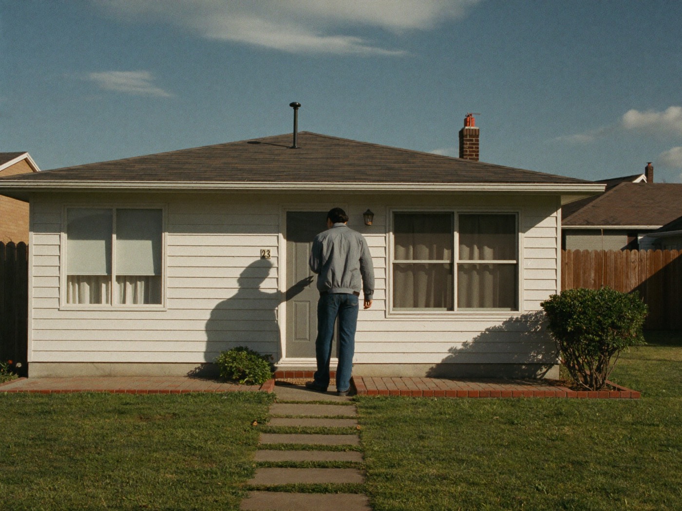 A man in a grey jacket knocking on the front door of a small single-storey house